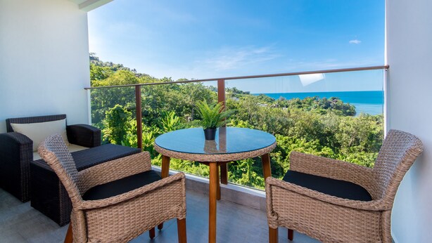 Ein Balkon mit Korbmöbeln, einem Glastisch und Blick auf eine üppige Landschaft und den Ozean.