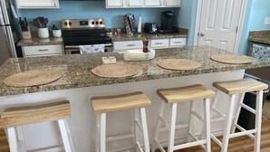 Een keuken met granieten aanrecht, een roestvrijstalen fornuis en witte kastjes. Er staan vier houten barkrukken en een koffiezetapparaat op het aanrecht.
