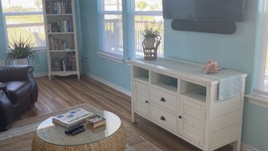 Een woonkamer met een ronde salontafel, een boekenkast, een aan de muur bevestigde televisie en een raam met jaloezieën.