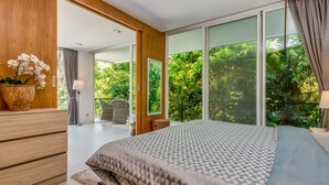 Een moderne slaapkamer met een groot bed, houten meubels en uitzicht op weelderige begroeiing door grote ramen.