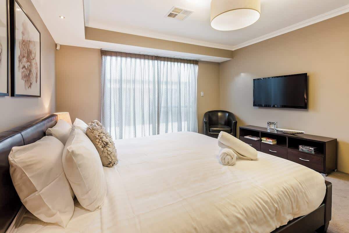 Un dormitorio con una cama grande, un televisor montado en la pared, una silla y una mesita de noche con una lámpara.