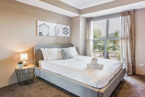 A bedroom with a large bed, a bedside table with a lamp, a wall clock, and a window with curtains.