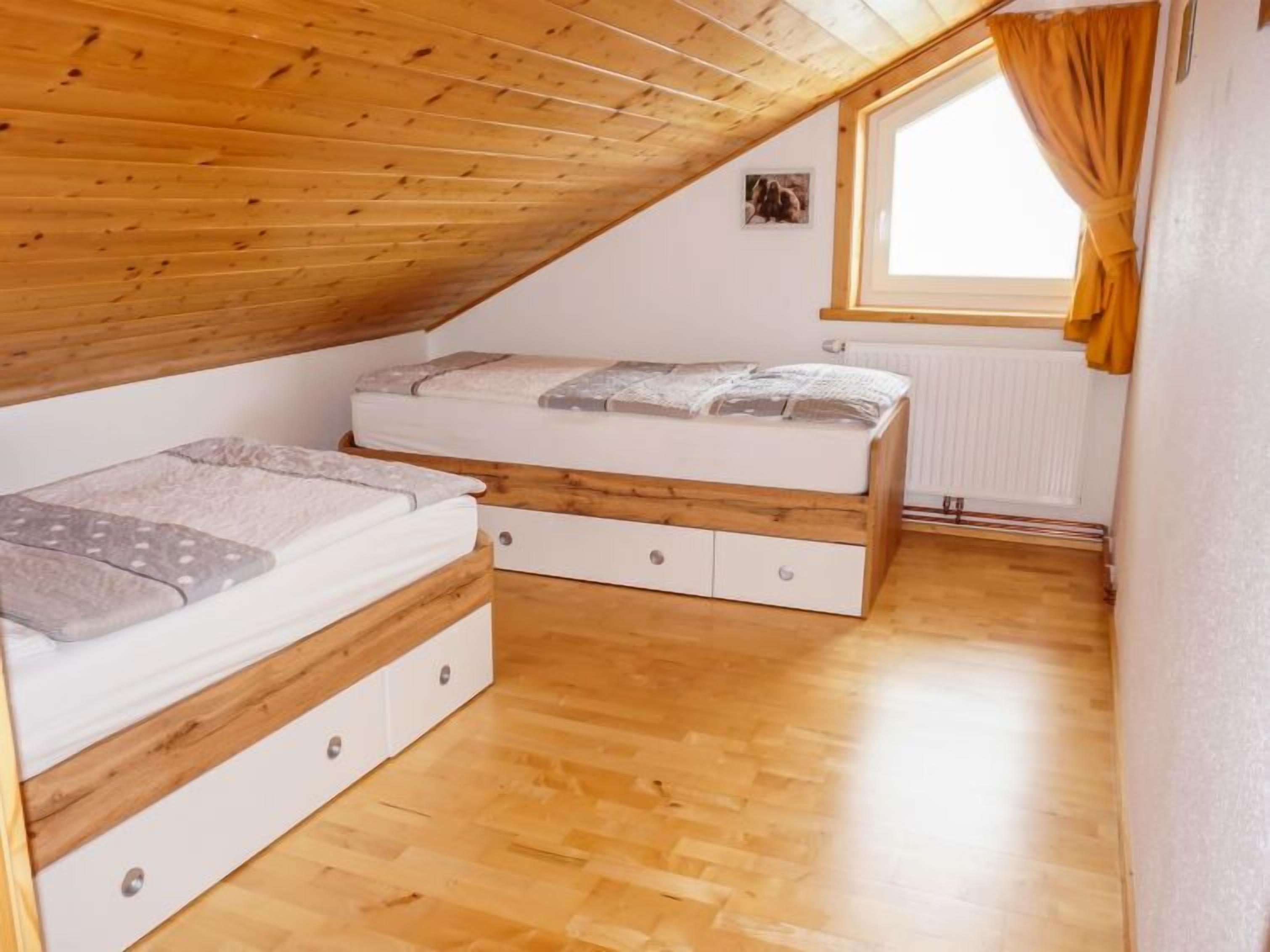 A small, wooden-paneled attic bedroom with two beds, a window with curtains, and a radiator.