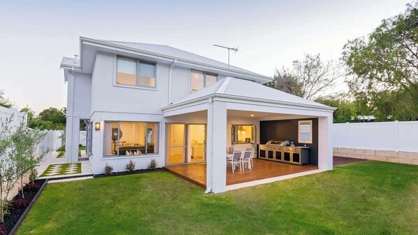 A modern two-story house with a covered patio, a visible kitchen area, and a well-maintained lawn.
