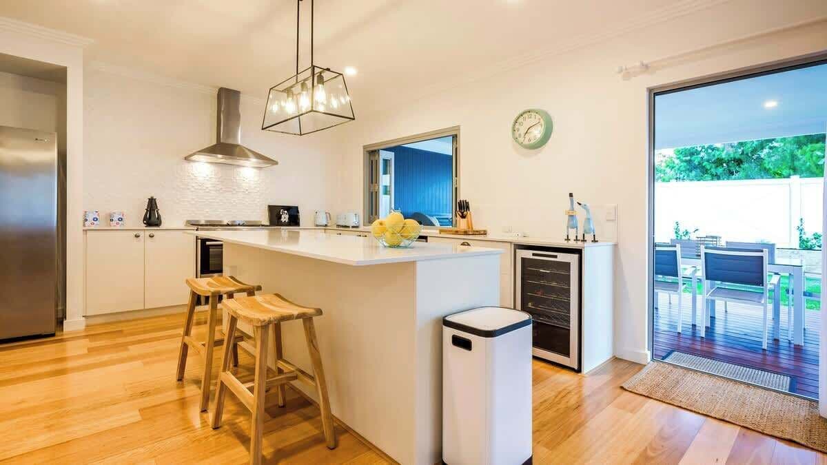 A modern kitchen with a central island, wooden stools, a stainless steel range hood, and a large window overlooking a deck.