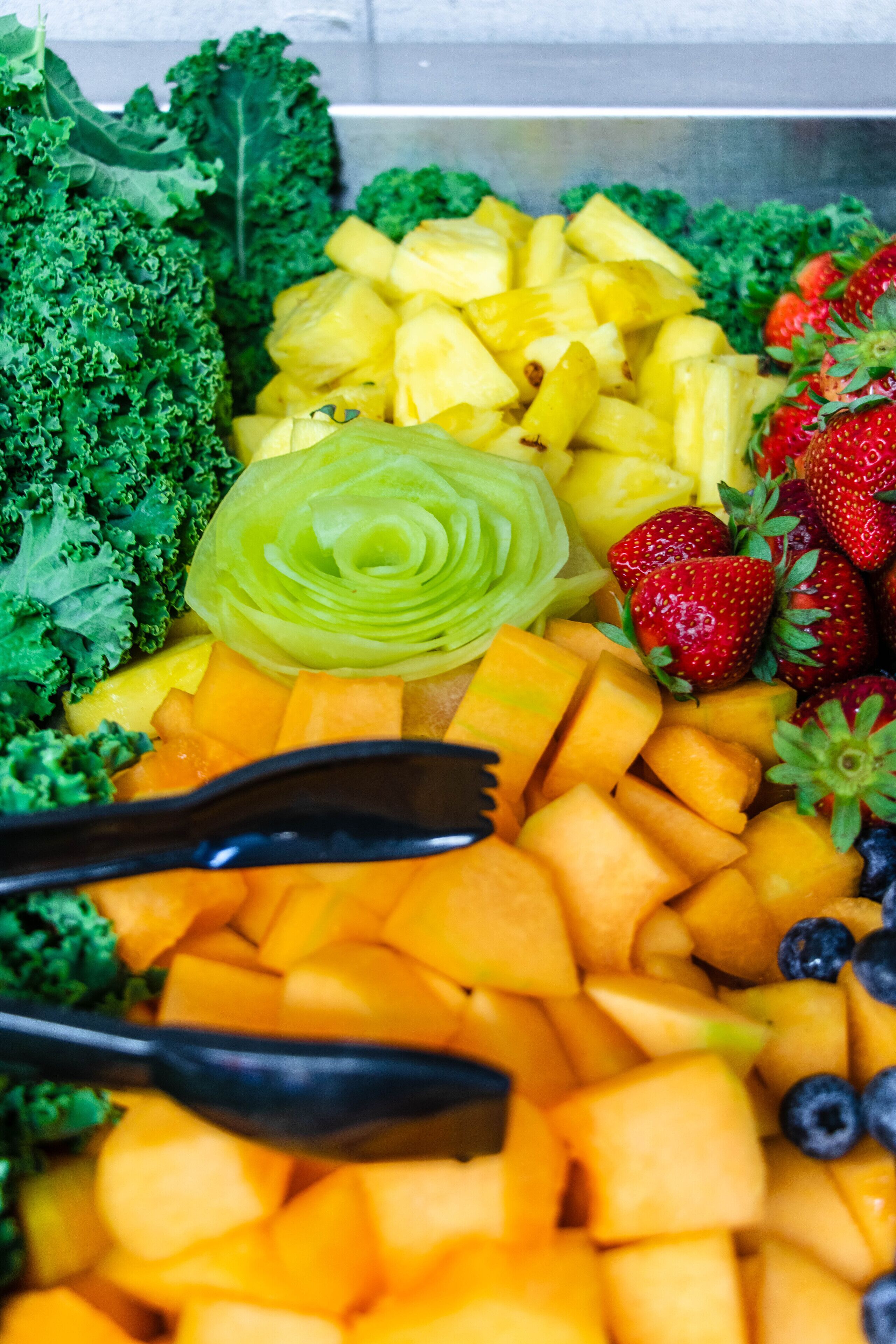 A variety of fresh fruits and vegetables including kale, pineapple, strawberries, and cantaloupe.