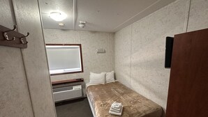Quarto pequeno e compacto com uma cama de solteiro, janela, aquecedor fixado na parede e alarme de incêndio.