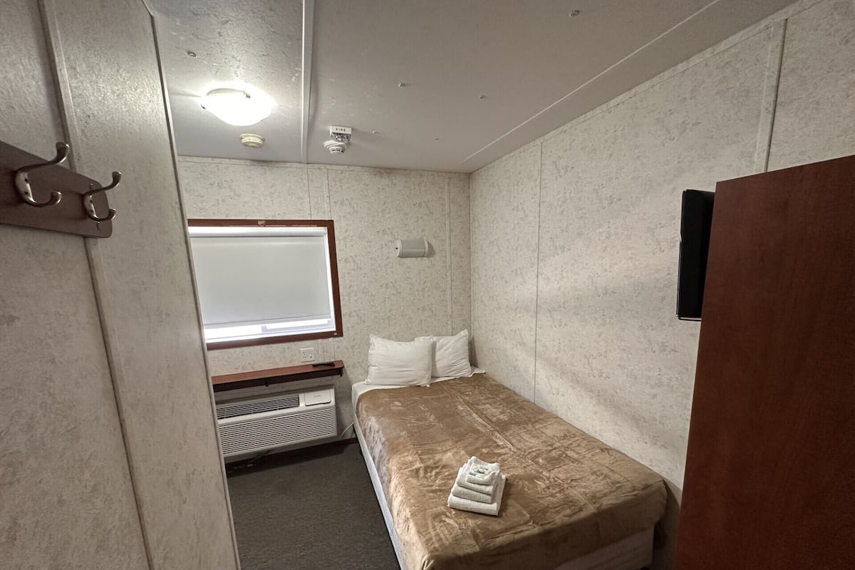A small, compact room with a single bed, a window, a wall-mounted heater, and a fire alarm.