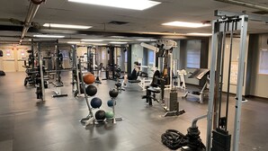 Uma academia com diversos equipamentos de ginástica, um suporte para pesos e uma pessoa utilizando uma máquina.