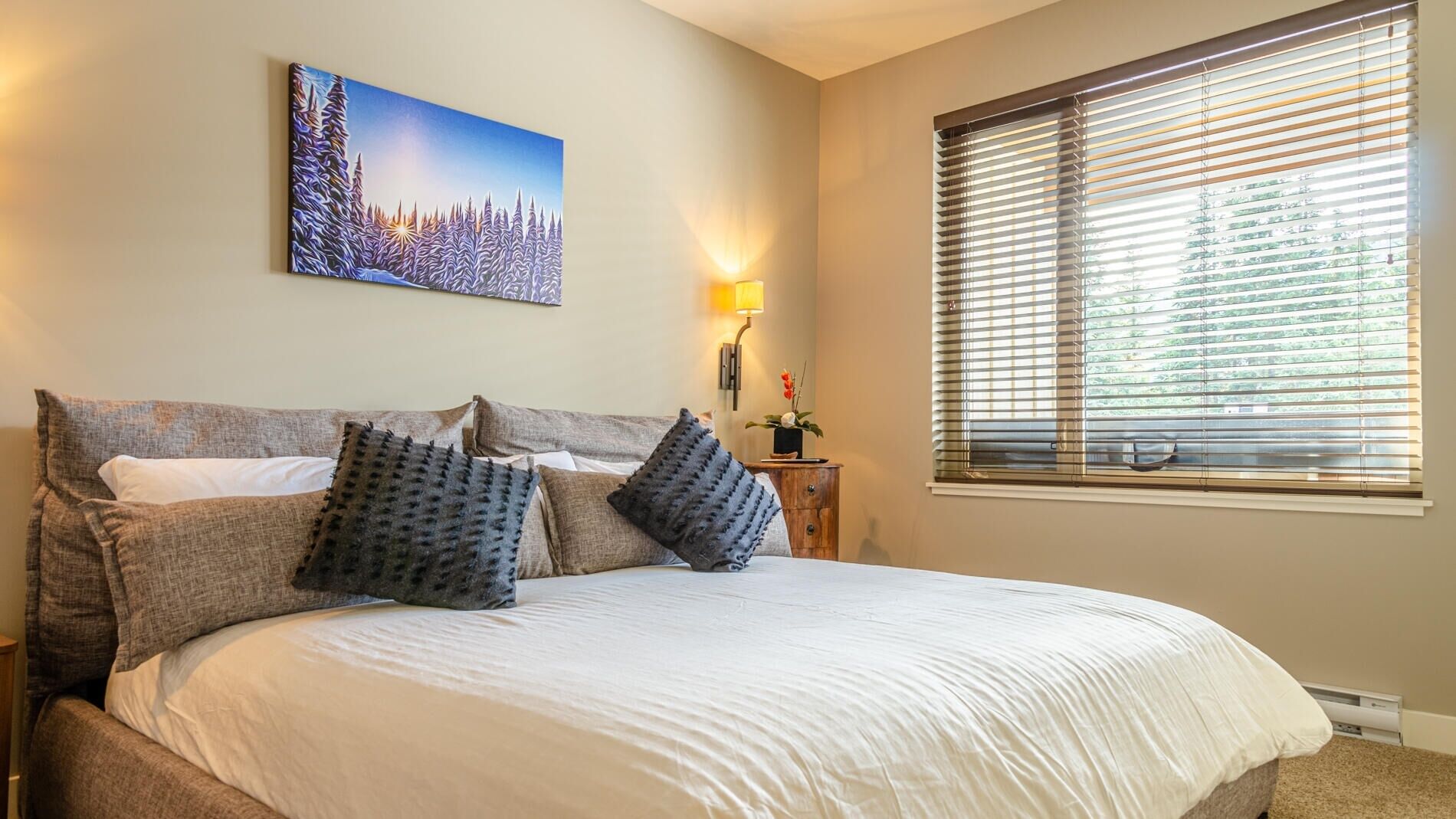 A bedroom with a large bed, a painting of a snowy landscape, a bedside lamp, and a window with blinds.