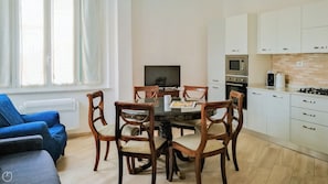 A modern kitchen with white cabinets, a microwave, oven, and a coffee maker. There is a dining table with chairs and a TV in the background. - Italianway - XX Settembre 55 (Alassio)
