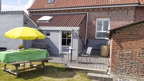 Een terras met een picknicktafel en een gele parasol, een bakstenen huis met een pannendak, en een kleine tuin.