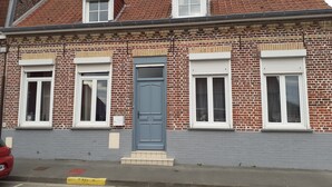 Een gebouw van baksteen met twee verdiepingen, voorzien van een rode dakbedekking, witte kozijnen en een blauwe deur.