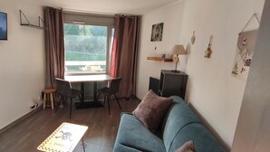 Ein Wohnzimmer mit einer Couch, einem Couchtisch, einem an der Wand befestigten Fernseher, einem Esstisch mit Stühlen und einem Fenster mit Vorhängen.