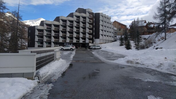Ein modernes Gebäude mit einer markanten, geometrischen Fassade, einem Parkplatz mit Schnee und einem Bergpanorama im Hintergrund.