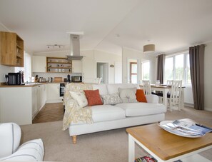 A modern, well-lit living room with a white sofa, a wooden coffee table, and a kitchen area with white cabinets and a built-in microwave. - Hollicarrs - Woodland Lodge (York)