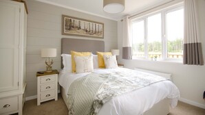 Een slaapkamer met een groot bed, een nachtkastje met een lamp, een lijst met een afbeelding aan de muur en een raam met gordijnen.