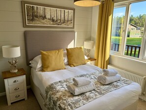 Ein ordentlich bezogenes Bett mit zwei Kissen, einer Nachttischlampe, einem Nachttisch mit Schubladen, einem gerahmten Waldgemälde über dem Bett und einem Fenster mit Vorhängen.