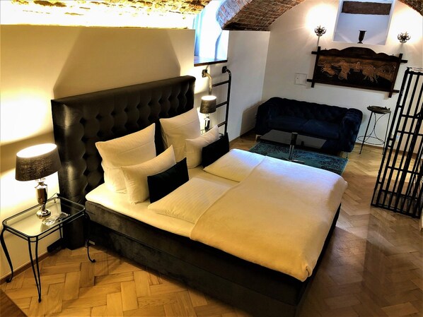 A bedroom with a large bed, a nightstand, a lamp, a sofa, and a wall-mounted shelf with a decorative item.