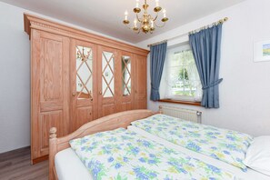 A bedroom with a wooden wardrobe, a bed with floral bedding, a window with blue curtains, and a chandelier.