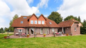 Een twee verdiepingen tellend bakstenen huis met een dak van rode tegels, een overdekte patio met buitenmeubilair en een verzorgd gazon.
