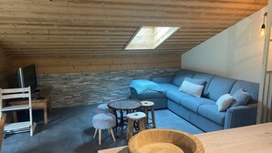 Ein gemütliches Dachzimmer mit schrägen Wänden, einer Steinwand, einem blauen Sofa, einem Holztisch und einem Fernseher auf einem Holzboden.
