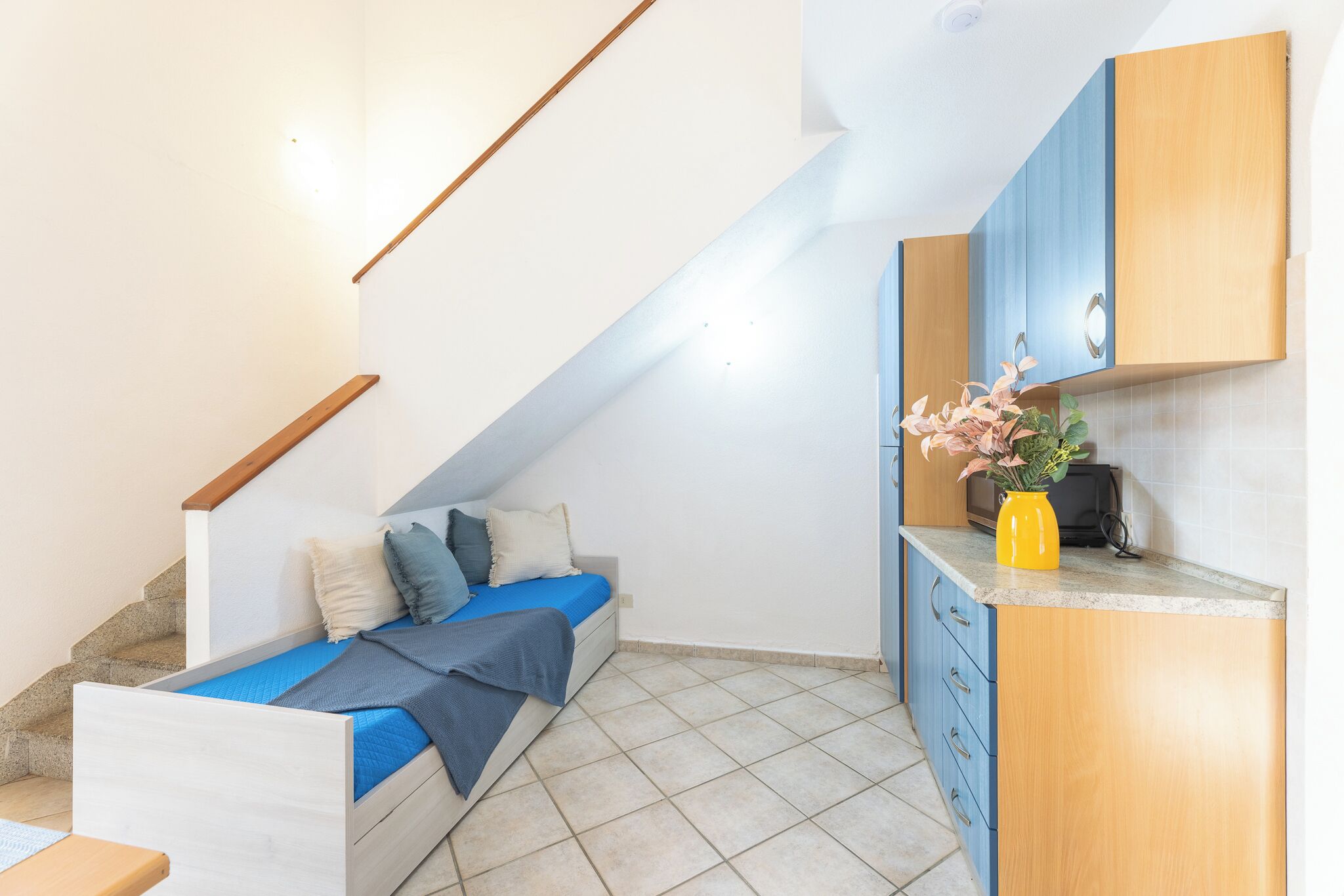 A bedroom with a bed, a small desk, a kitchen area with cabinets, and a staircase.