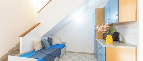 A bedroom with a bed, a small desk, a kitchen area with cabinets, and a staircase.