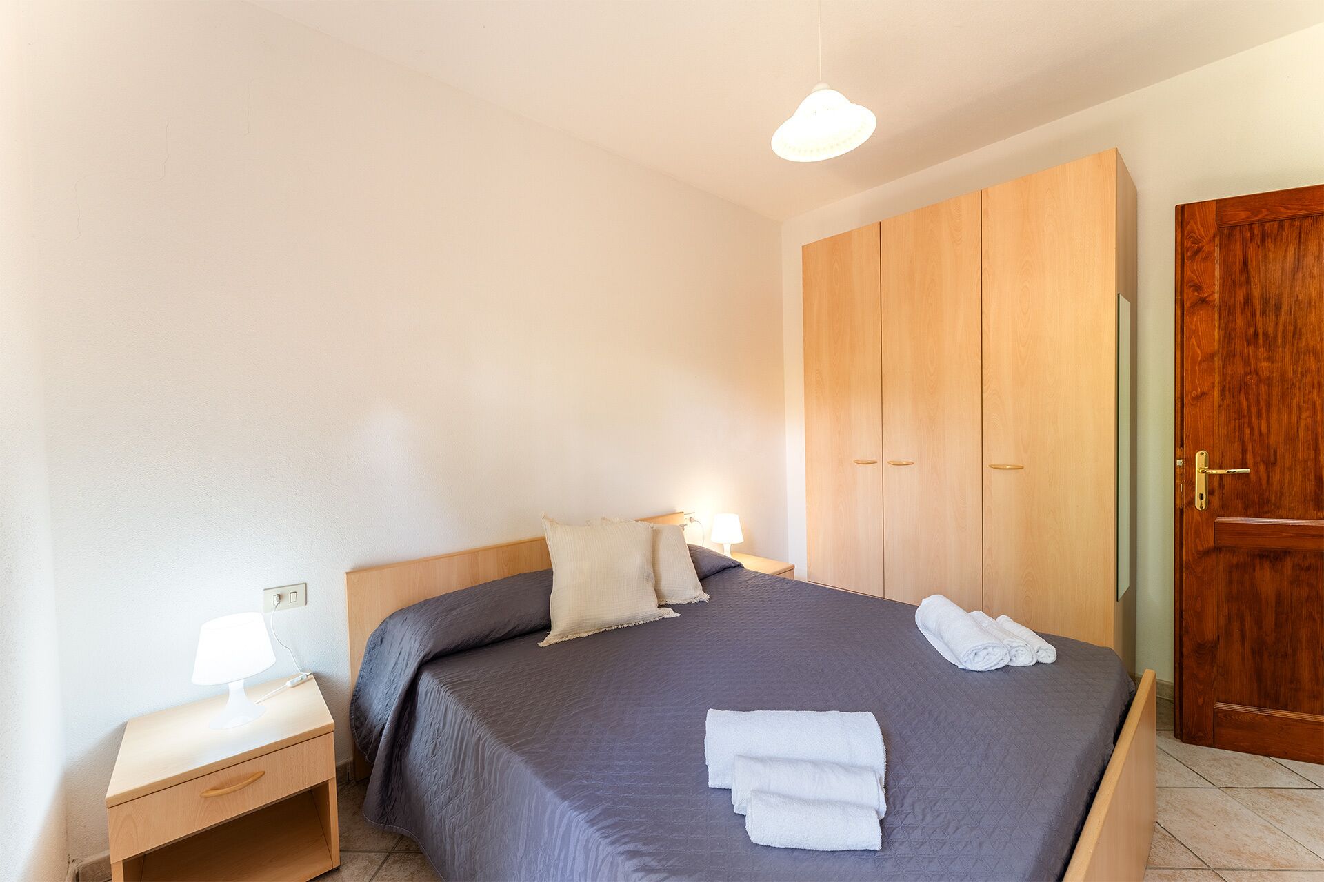 Quarto com cama, mesa de cabeceira, luminária e guarda-roupa de madeira.