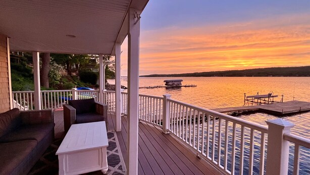 A covered deck with outdoor furniture, a dock extending into a lake, and a sunset in the background.