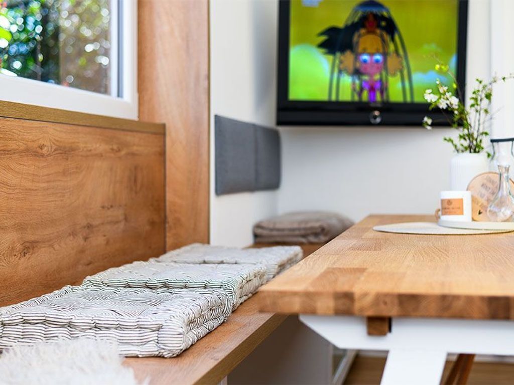 Eine Holzbank mit einer gestreiften Decke, ein Holztisch mit einer weißen Tischmatte, ein an der Wand befestigtes Fernsehgerät und eine Vase mit Blumen auf dem Tisch.