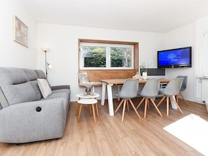 Ein modernes Wohnzimmer mit einem grauen Sofa, einem Esstisch mit Stühlen, einem Fernseher und einem Fenster mit Blick auf Grünflächen.