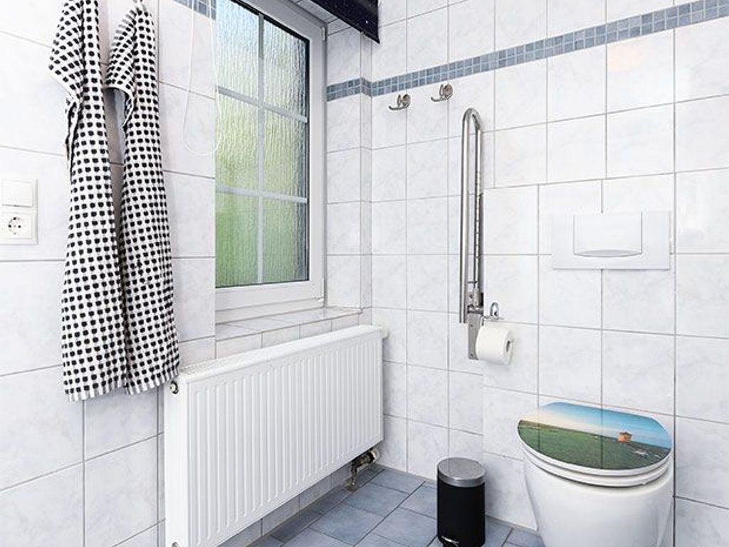 Een badkamer met witte tegels, een raam, een toilet met een groen landschapsdesign en een zwarte prullenbak.
