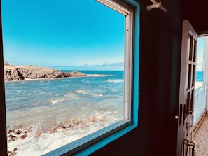 Une vue par la fenêtre sur une côte rocheuse, où les vagues s’écrasent contre le rivage.