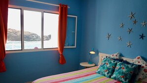 A bedroom with a bed, a window with curtains, a nightstand, and starfish wall decor. - Casa Azul Holiday Home (El Cotillo)