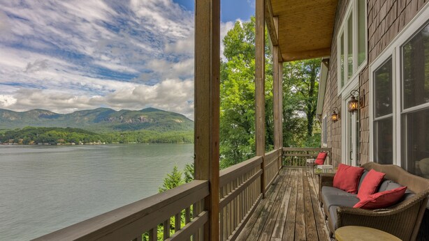Eine Holzterrasse mit Blick auf einen See und Berge. Eine Rattan-Couch mit roten Kissen und ein kleiner Tisch sind vorhanden.