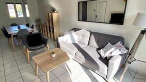 A living room with a sofa, a coffee table, a dining area with chairs, a bookshelf, and a mirror. - Villa proche de la mer (Concarneau)