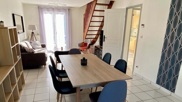 A dining area with a wooden table and blue chairs, a staircase, a sofa, and a bookshelf. - Villa proche de la mer (Concarneau)