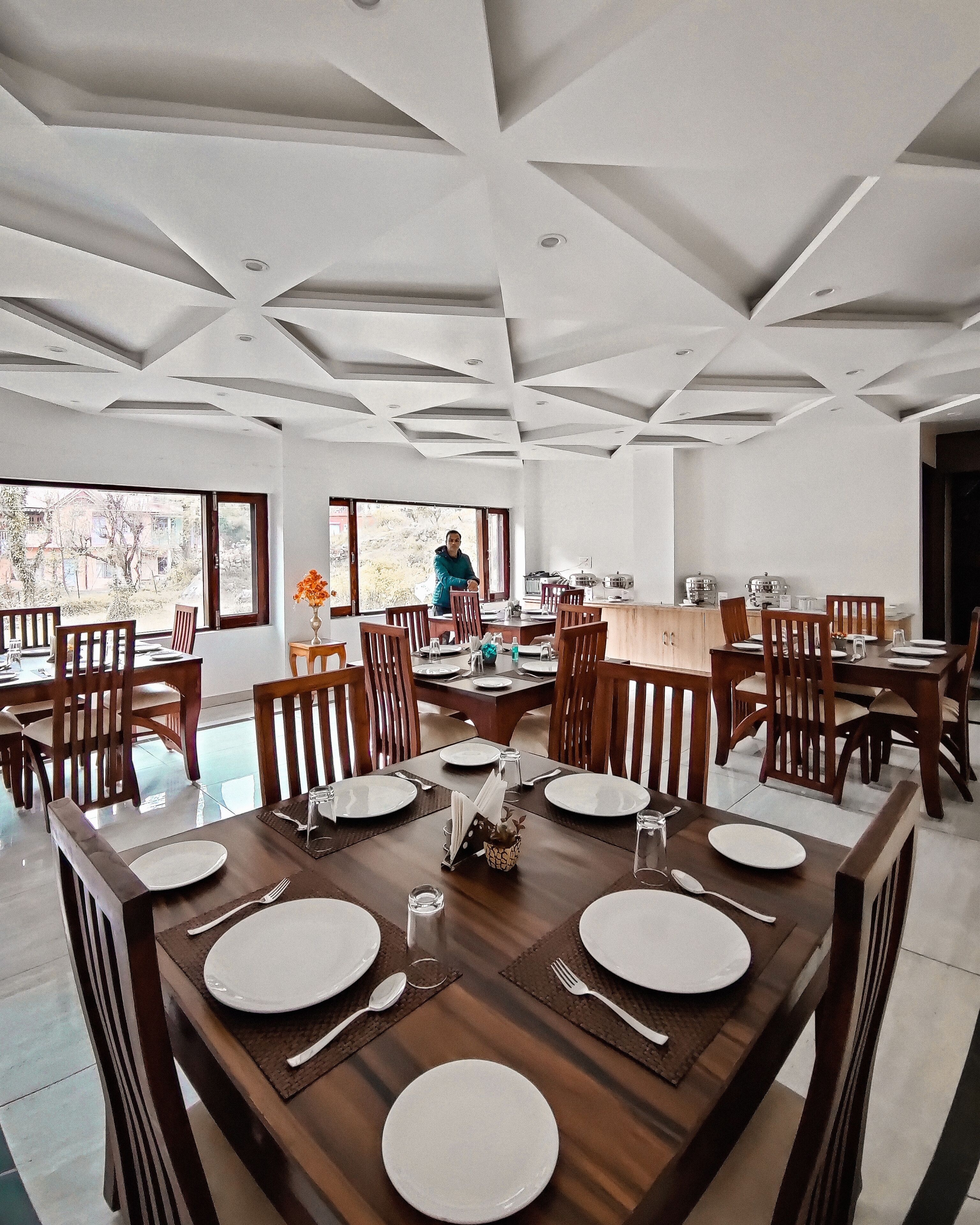 A dining area with a high ceiling and large windows, featuring a central round table with chairs and a person in the background.