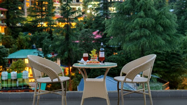 A wicker table with two chairs, a bottle, and two glasses of red wine on a balcony overlooking a cityscape with buildings and trees. - Golden Rock - AM Hotel Kollection (Dharamshala)