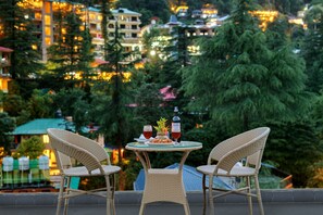 A wicker table with two chairs, a bottle, and two glasses of red wine on a balcony overlooking a cityscape with buildings and trees. - Golden Rock - AM Hotel Kollection (Dharamshala)