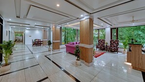 A spacious lobby with a modern design, featuring a reception desk, seating area with a pink sofa, and large windows offering a view of greenery outside. - Golden Rock - AM Hotel Kollection (Dharamshala)