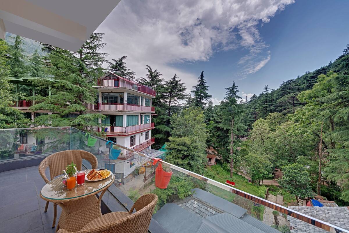 A balcony with wicker furniture, a dining table, and a glass railing overlooking a forested area and a building with a red balcony.