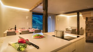 A modern kitchen with a large island, a dining area with a table set for two, and a view of a landscape through a window. - Chalet 'Ferienhaus Im Stadl' with Mountain Views, Private Terrace and Wi-Fi (St. Martin in Passeier)