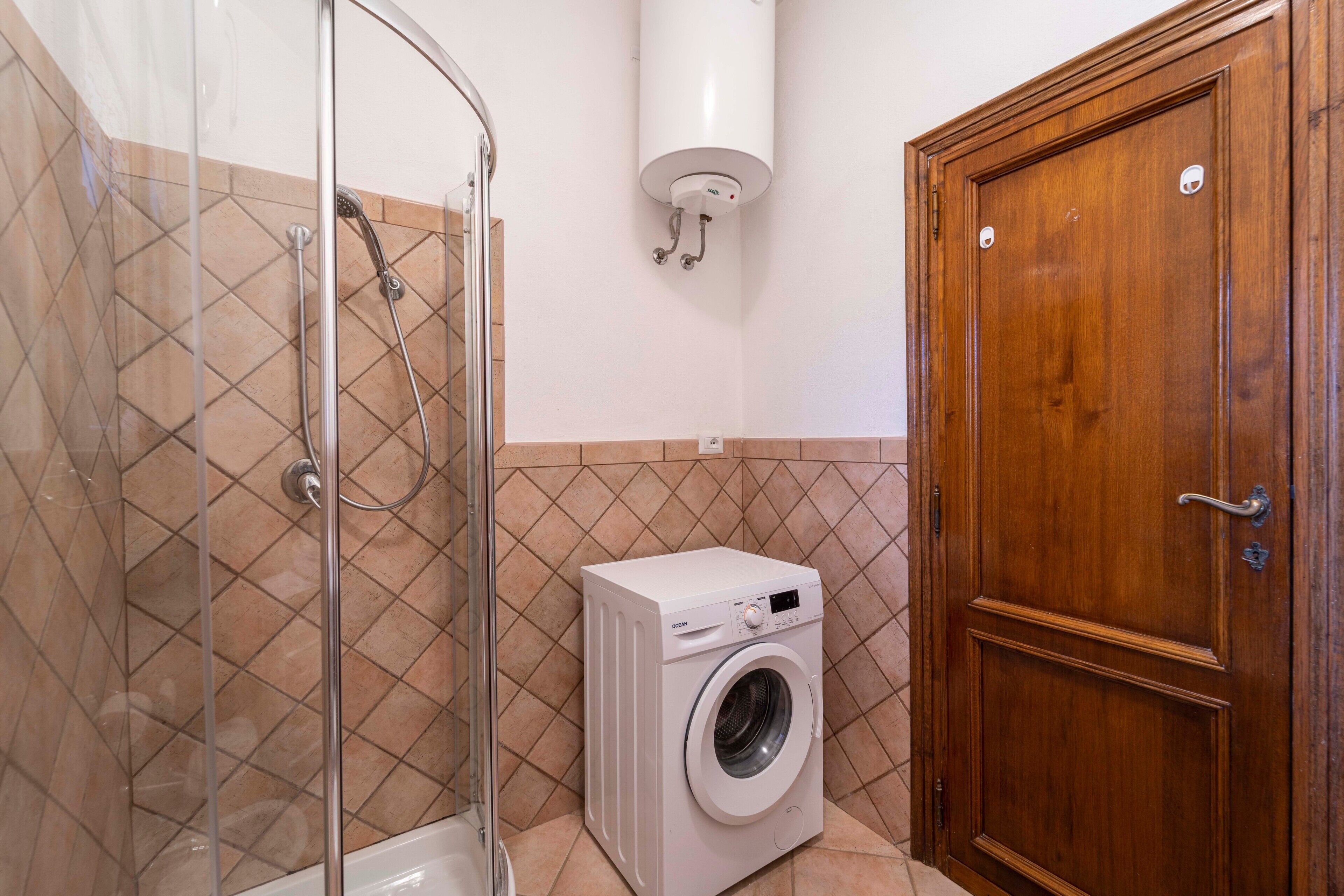 A bathroom with a glass shower enclosure, a washing machine, and a wooden door.