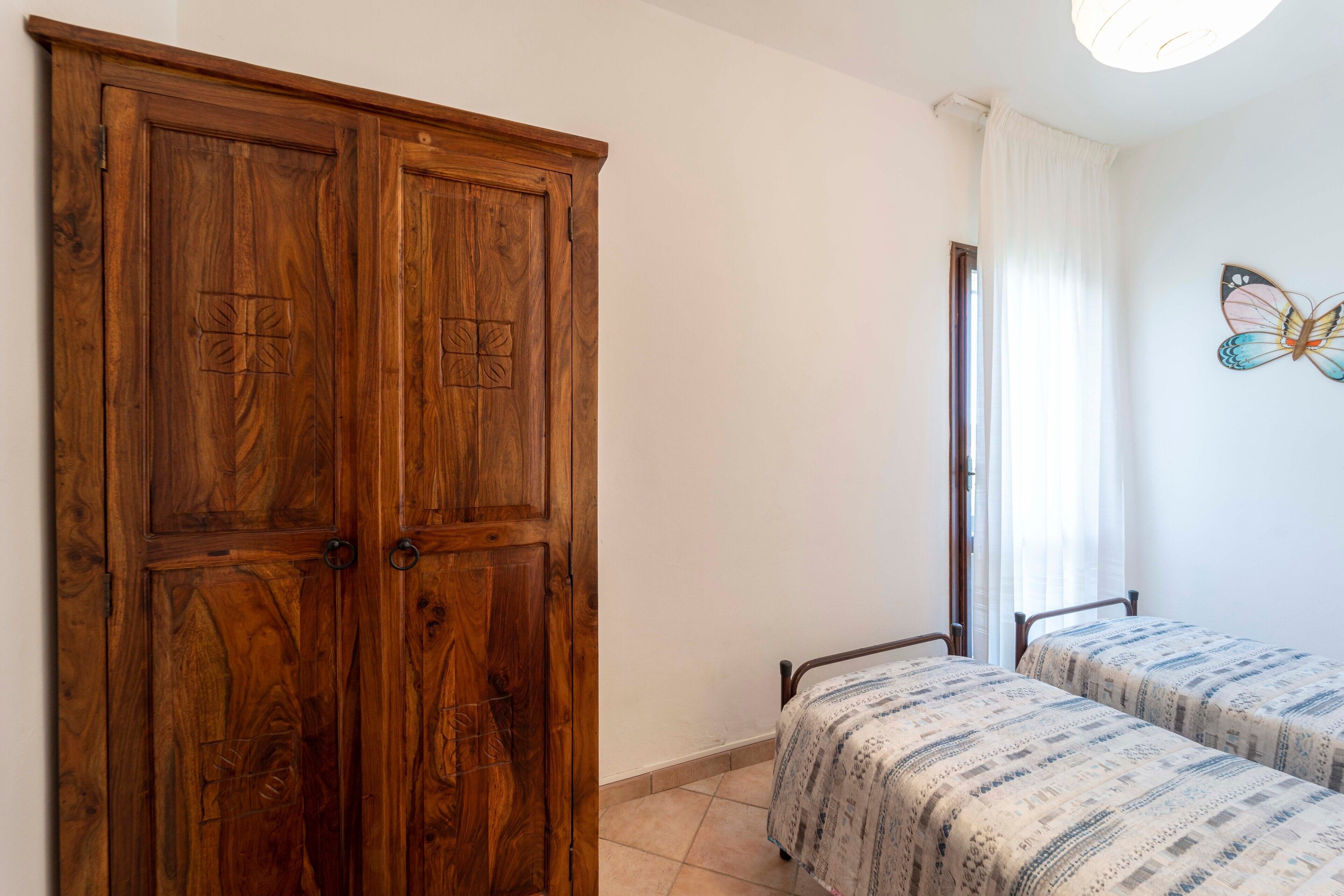 Een slaapkamer met een houten garderobe, twee bedden met beddengoed met een patroon, een raam met gordijnen en een vlinder decoratie aan de muur.