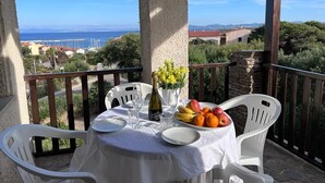 Una mesa con mantel blanco, sillas blancas, una botella, copas y un frutero en un balcón con vistas al mar y edificios.