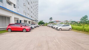 A parking lot with multiple cars parked in rows, a building with balconies, and a palm tree in the background. - Life 2837 Apartment Dramaga Tower (Dramaga)