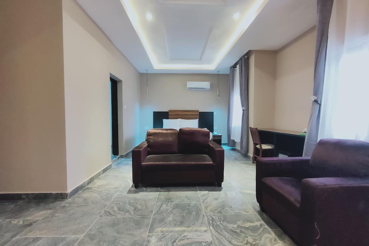 A spacious living area with a brown leather sofa, a purple armchair, and a television. The room has a tiled floor and a ceiling with recessed lighting.