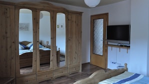 A bedroom with a wooden wardrobe, a bed with a blue and white patterned bedspread, a television on a stand, and a white ceiling light.